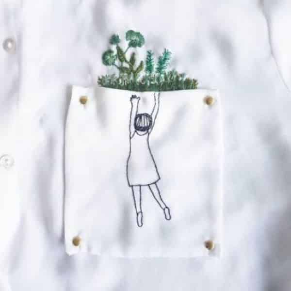 desenho de menina pendurada na beirada do bolso da camisa com jardins bordado dentro