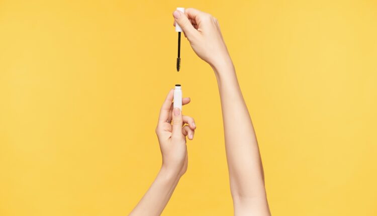 Indoor shot of young pretty woman’s hands with nude manicure being raised while holding bottle with mascara, going to dye eyelashes while being isolated over orange background